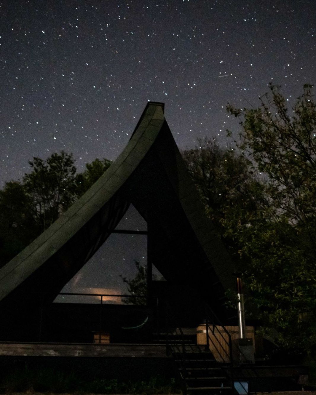 Un écolodge en A sous un ciel étoilé, une lumière allumée derrière la baie vitrée
